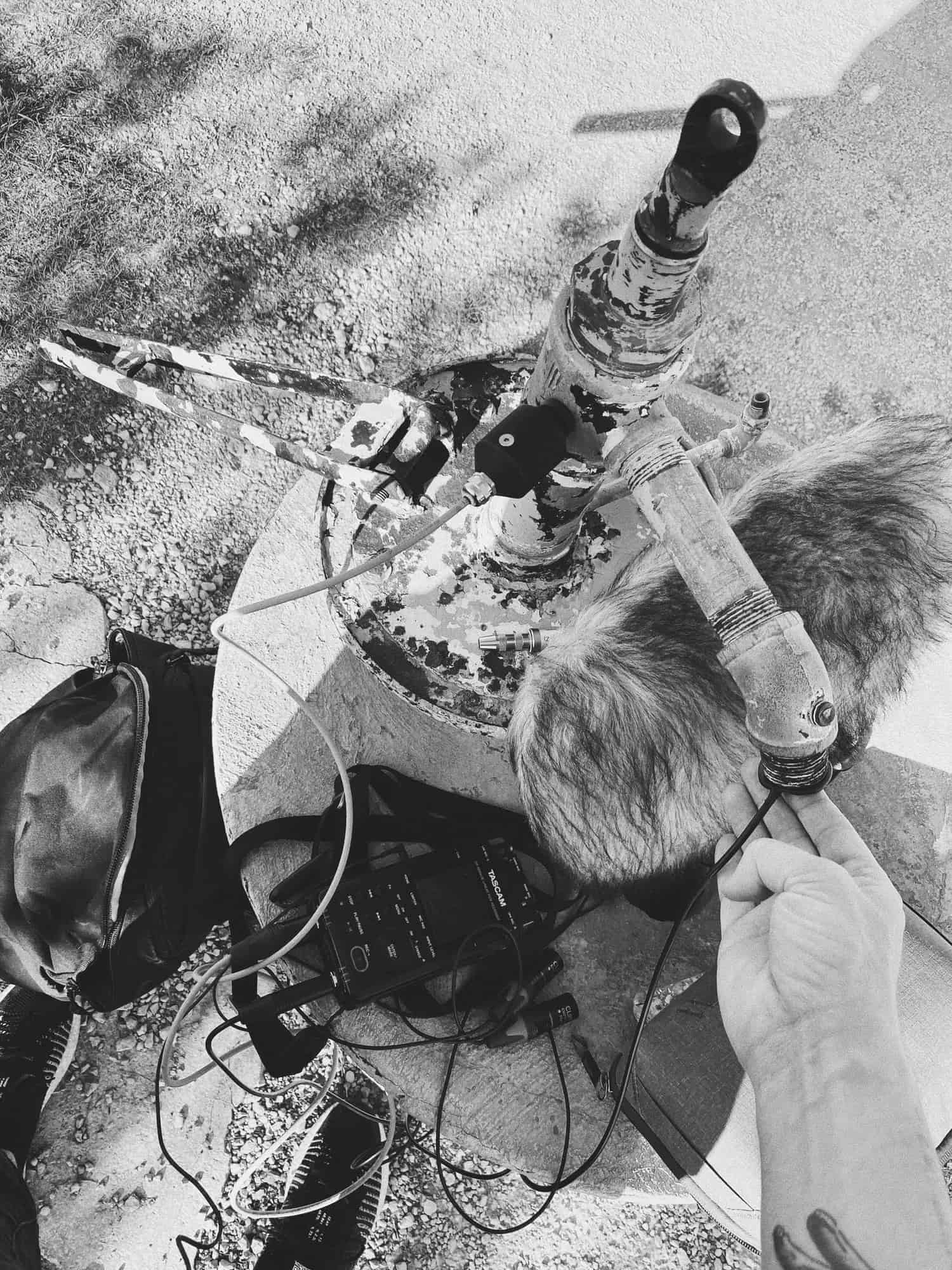 Black and white photograph of a hand holding a contact microphone against an old rusted water pump.